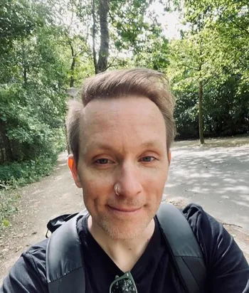 I am looking at the camera and smiling. I am wearing a black t-shirt and the straps of my black backpack are visible. In the background are sunny green trees and a walking path.